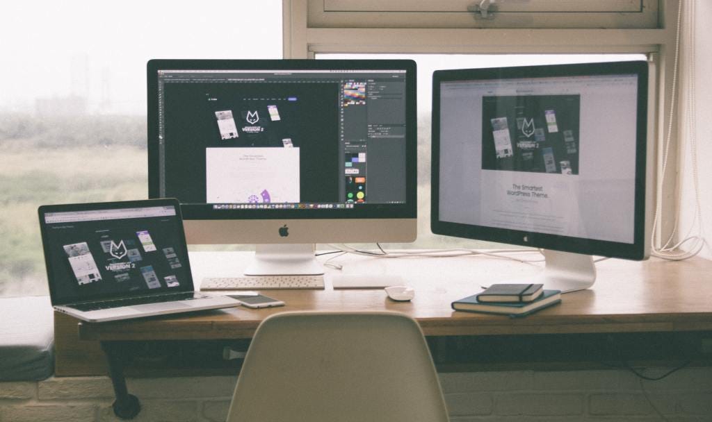 Two desktop computers sit on a desk with a laptop left on the desk