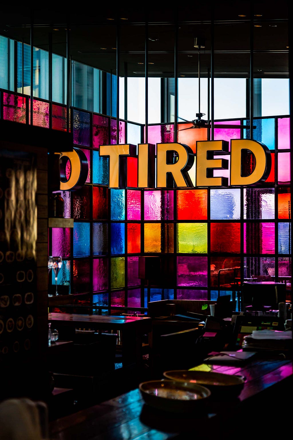 A big sign that spells out the word tired in all capitalized orangeletters in front of a rainbow or multicolored glass wall. There are chairs in front of both.