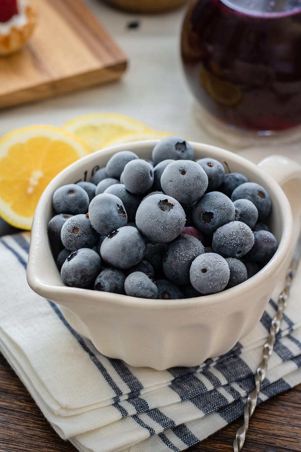 A bowl of frozen blueberries sitting on a blue and white kitchen towel with lemons and blueberry jam in the background.