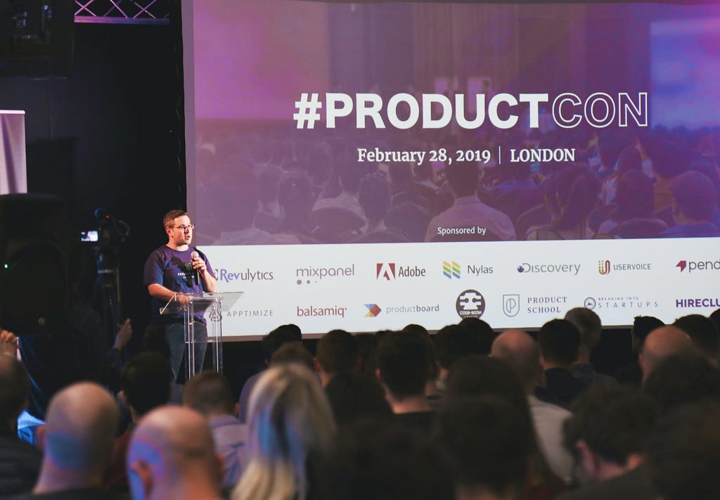 A speaker stands in front of an audience. Behind the speaker is a presentation titled #productcon.