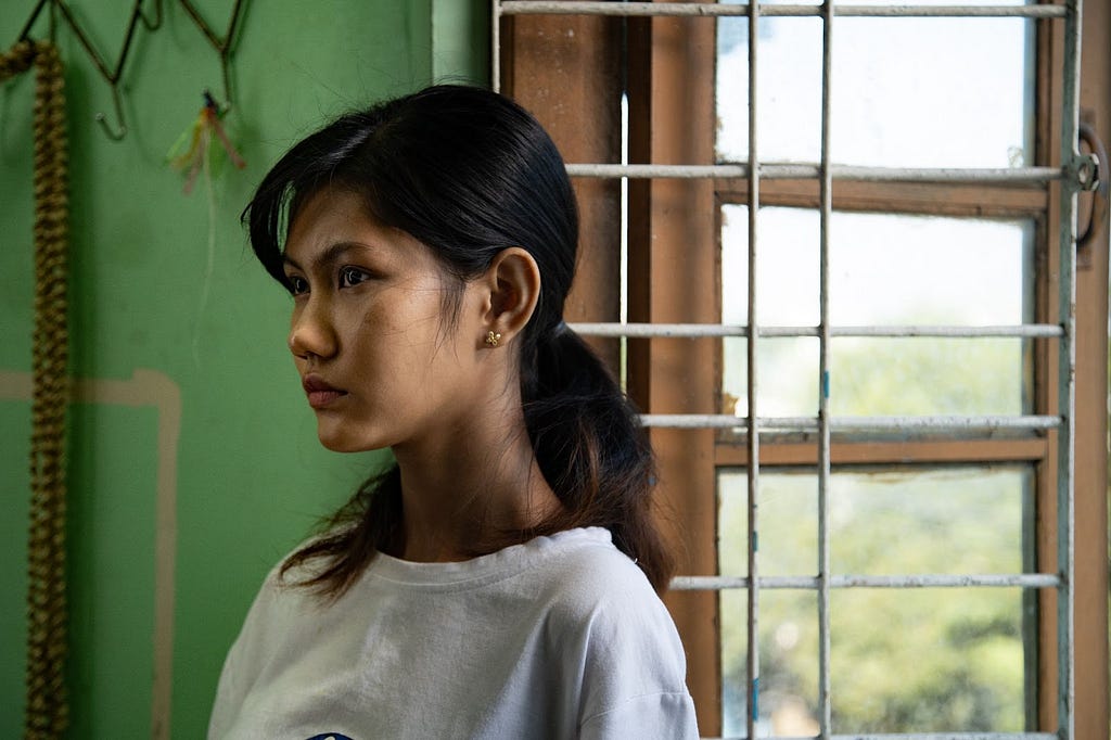 San is leaned against her window sill, framed on her left by a green wall and on the right by a wooden window enforced with metal bars. She is staring grimly into her home.