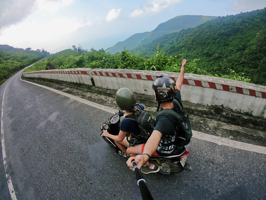 Two people riding a scooter away from the camera on a mountain road. The photograph appears to have been taken by the pillion passenger who can be seen holding a selfie stick.