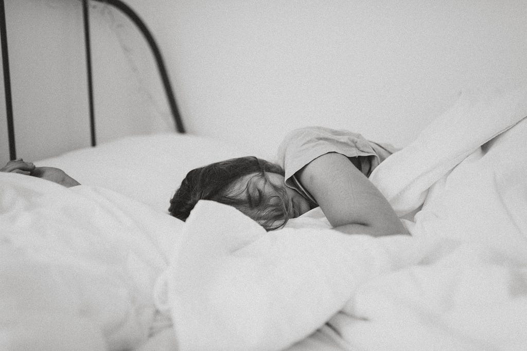 Black and white image of a girl laying in bed. She’s covered with white blankets, with one arm and shoulder over the covers, and her face partly showing. Her brown hair partly covers her face, and her eyes are closed.