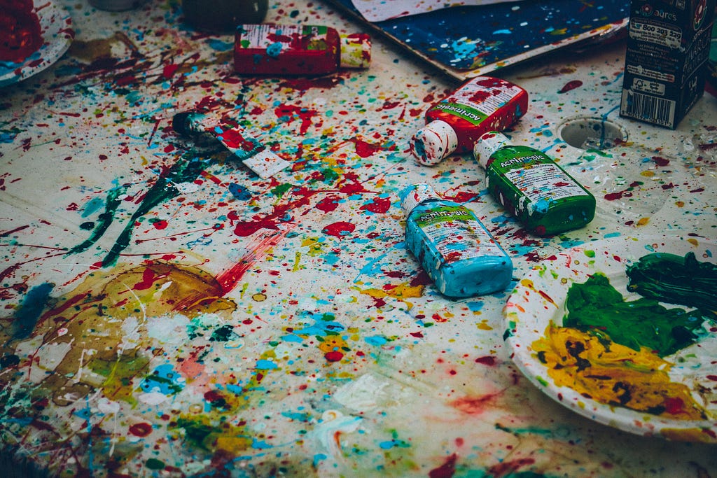 A colorful mess of paint splatters, with paint bottles, a dish and a laptop