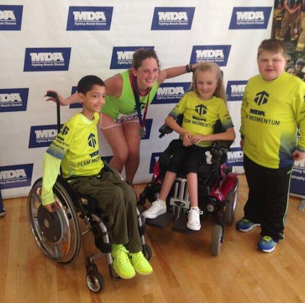 A photograph of Megan wearing a medal after she completed the Chicago Marathon. Megan is pictured with two children in wheelchairs and another standing who are all wearing bright yellow Muscular Dystrophy Association shirts.