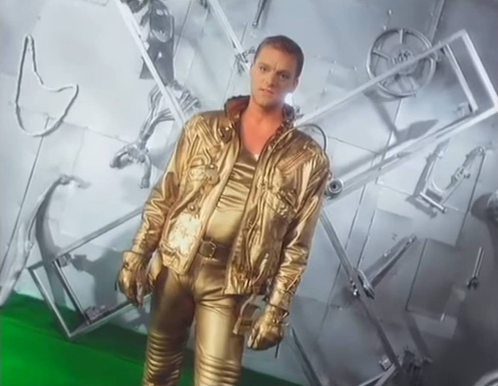 Andy Bell, vocalista do duo Erasure, posa em roupas douradas no clipe de seu cover de Lay ALl Your Love On Me.