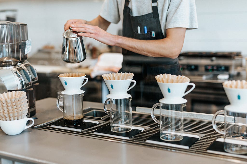 A Blue Bottle Coffee bar. Four glass containers with handles stand atop weighing scales. They have Blue Bottle brand filters on top. A barista pours hot water from a silver kettle into the first of the four filters. Dark brown coffee drips into the glass container.
