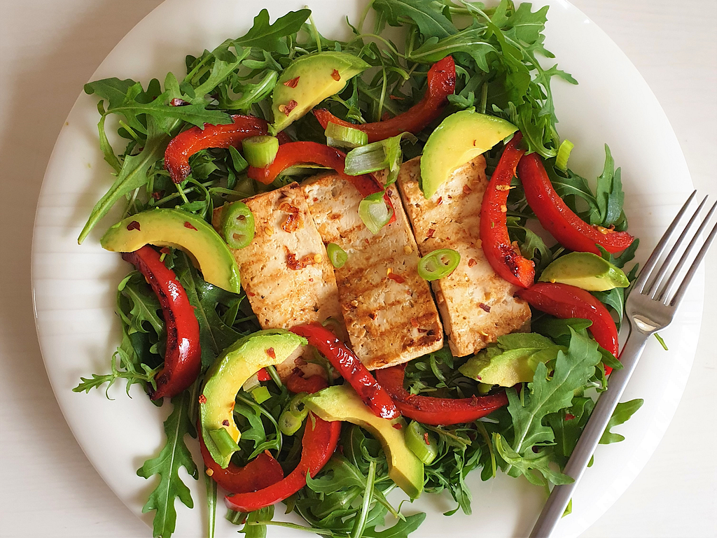 White plate with salad with green rocket leaves, red peppers, avocado and tofu