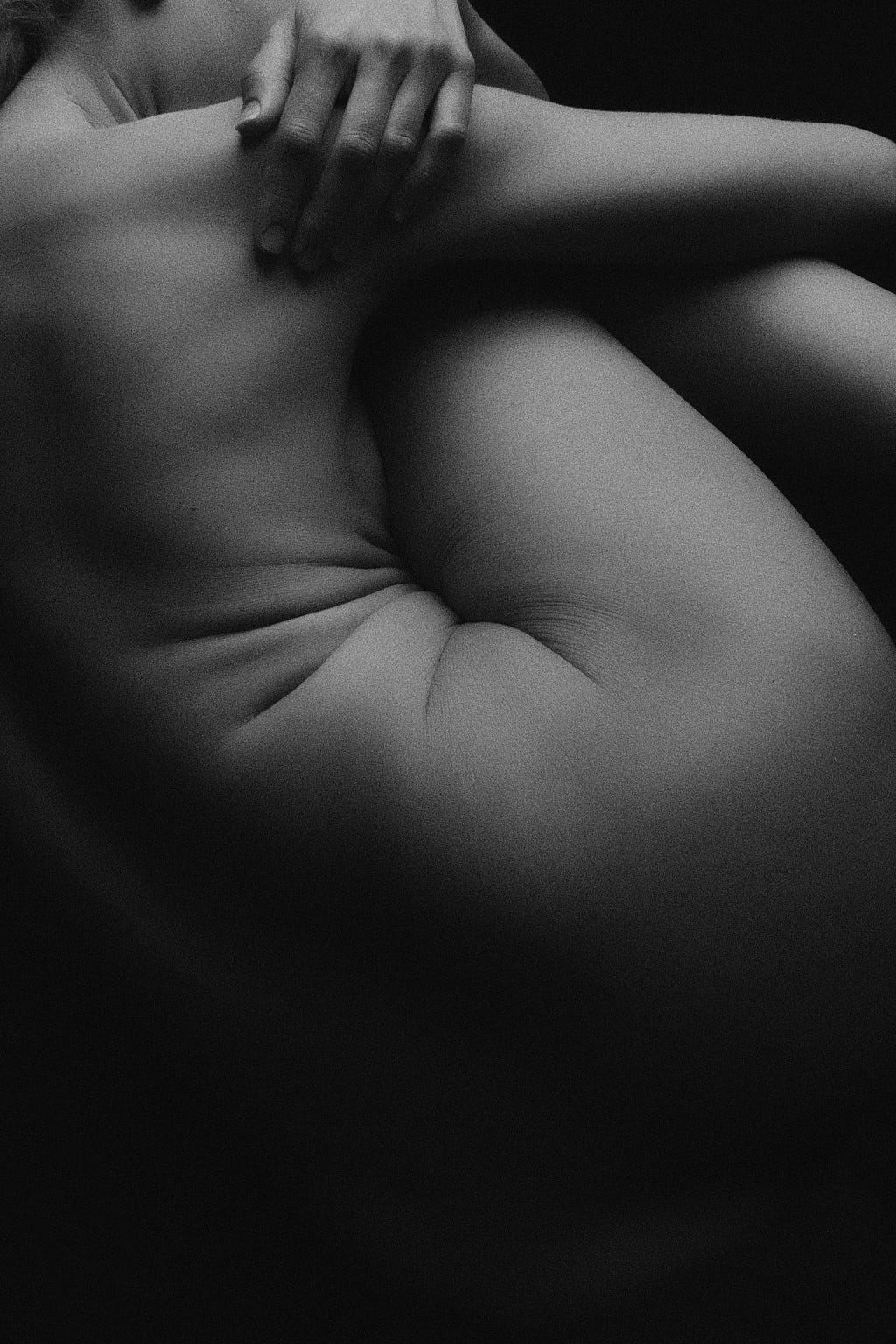 A black-and-white image of a woman’s torso and upper leg. The photograph is taken from a back and side profile perspective. The focus of the image is the folds on the woman’s body.