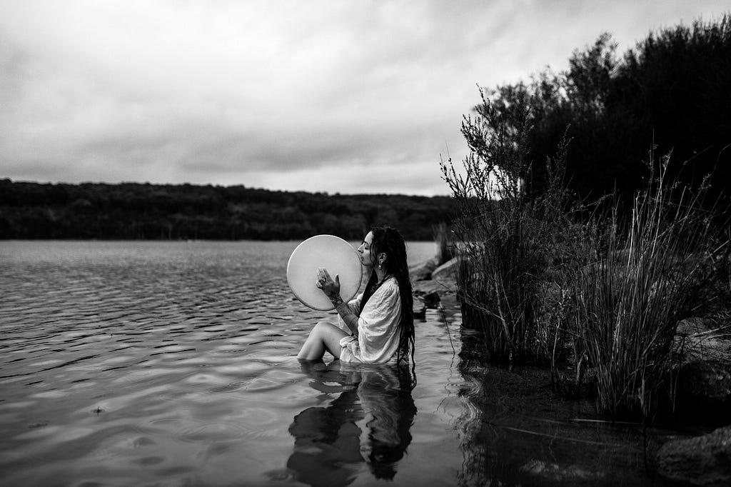 A person with long dark dreadlocks and a flowing white garment sits in a river or sea with tree-ed land in the distance, knees up, holding a large bodhran type drum.