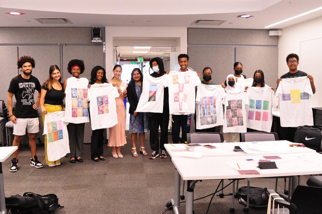 Group photo of Cambridge Mayor Sumbul Siddiqui with Cynthia Breazeal, Raechel Walker, 7 Cambridge Rindge and Latin School students, and 3 undergraduate researchers holding up t-shirts the students made.