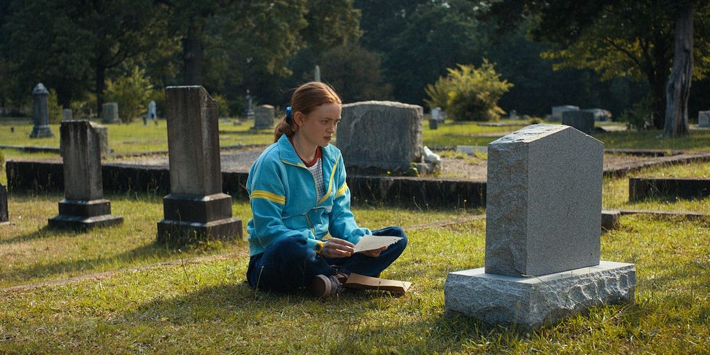 A still from the episode featuring Max, a young teenage girl with red hair pulled back in a ponytail and a blue windbreaker, sitting cross-legged in front of a headstone, holding a piece of paper in her hand.