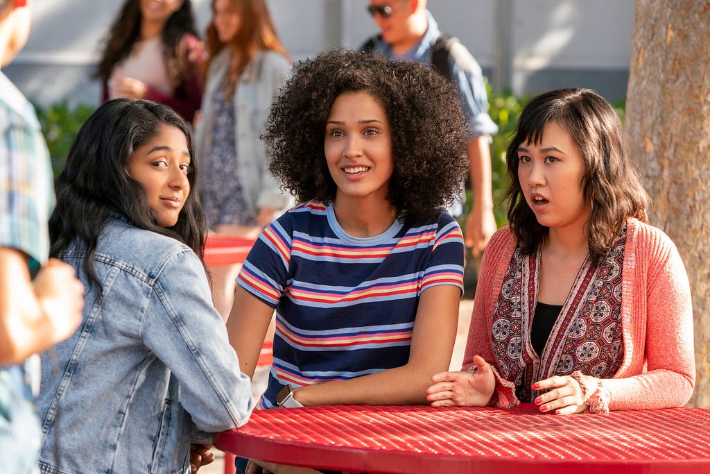 a screencap from the sitcom Never Have I Ever, depicting main characters, (left to right) Devi, Fabiola, and Eleanor, sitting at a table outside.