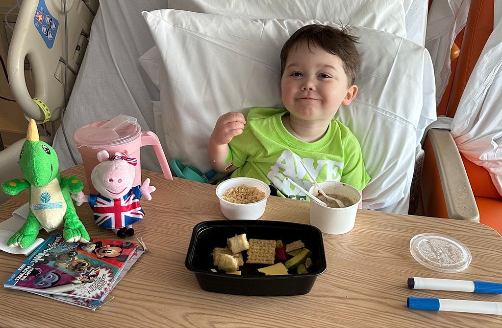 Child smiling in hospital bed with snacks and toys in front of him