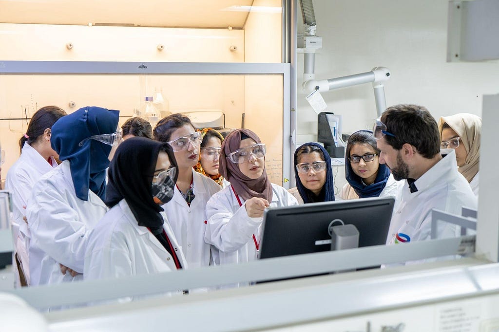 Young women wearing lab coats gather around a computer screen