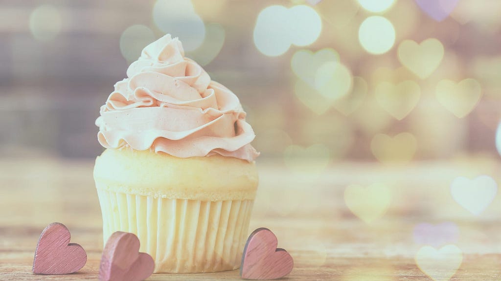 A vanilla cupcake with pink frosting sits beside pink wood hearts.