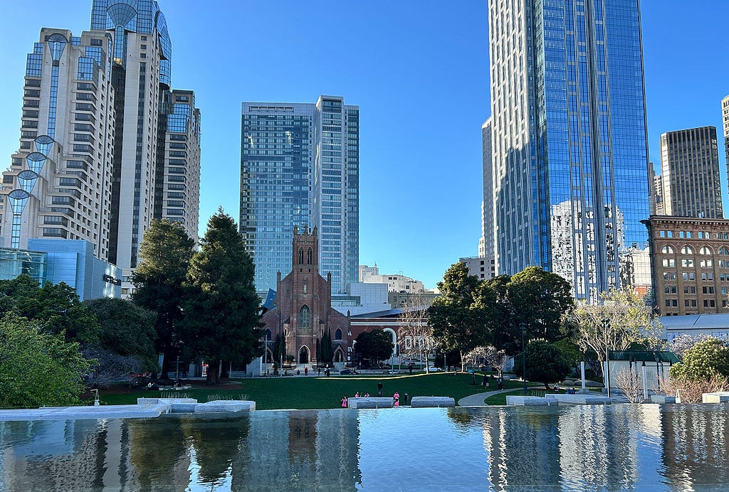 A pool of water overlooking San Francisco’s Yerba Buena Gardens. The blue sky and surrounding buildings reflect perfectly in the water, creating a mirror effect.