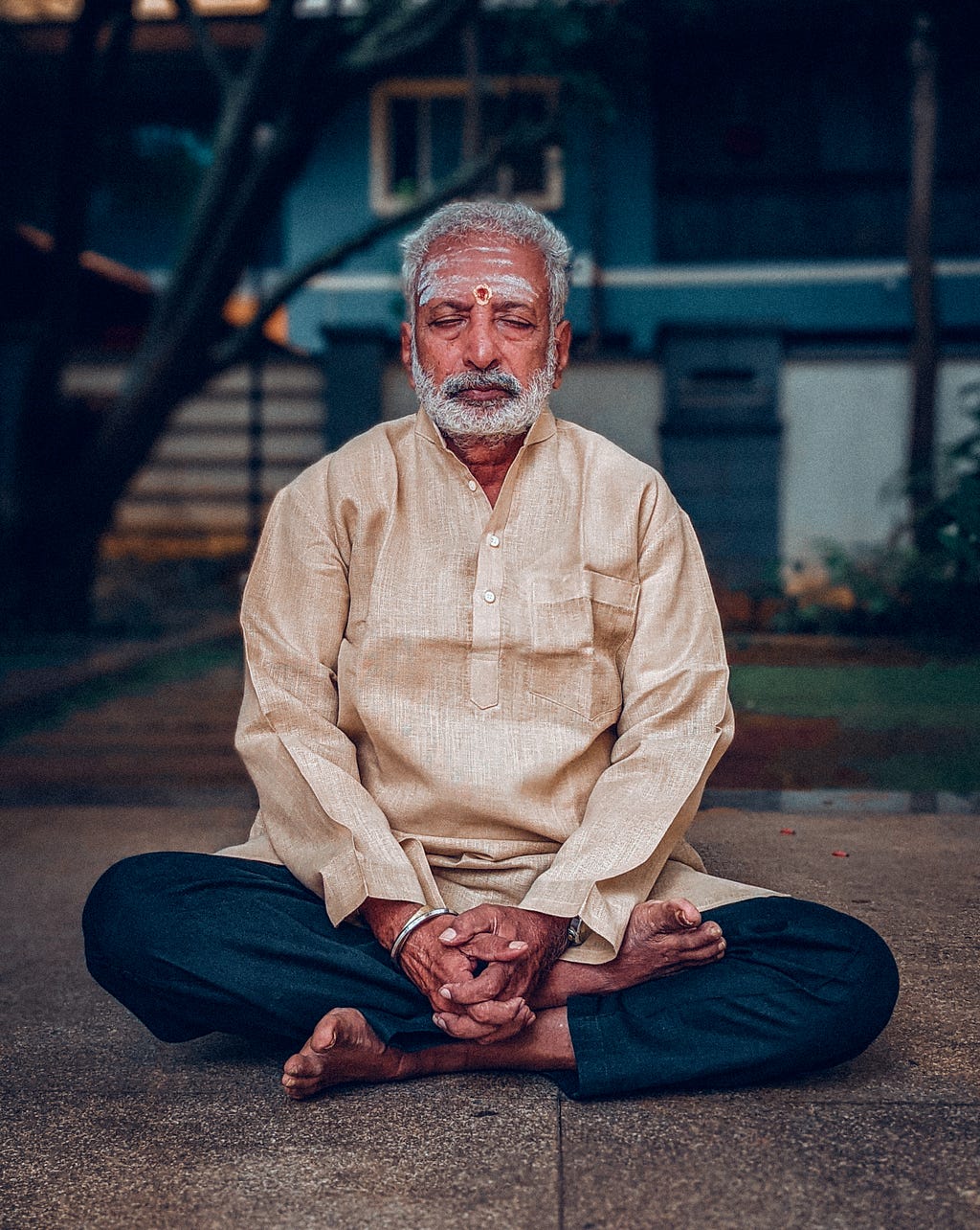 A man sitting on the floor cross legged with hands folded at his feet, eyes closed, meditating.