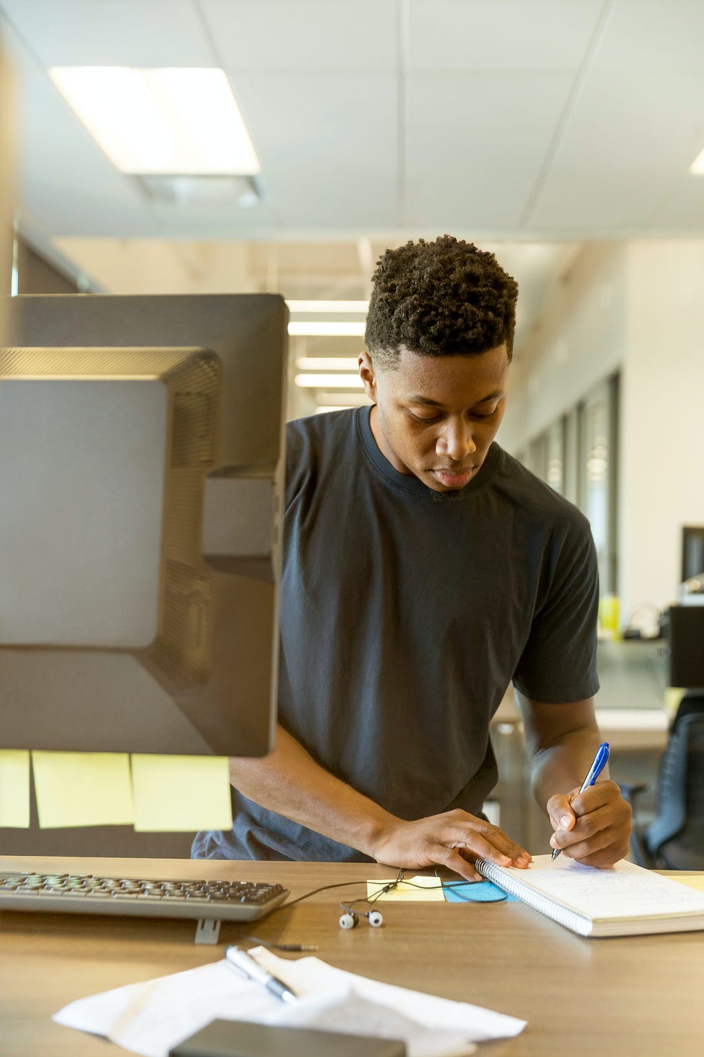 modern office setting. strip lighting, bland everything. a young man stood in front of a computer writes on a pad of paper to his left (camera right)