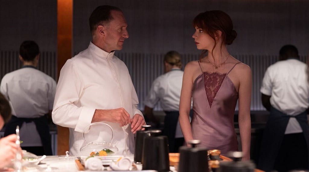 An image shows actress Anya Taylor-Joy in her character of Margot from The Menu. She wears a pink dress and her hair is an in updo. She looks surprised and to her right at Ralph Fiennes, who is in character from the movie. He is in chef’s attire. There are multiple sous-chefs working in the background.