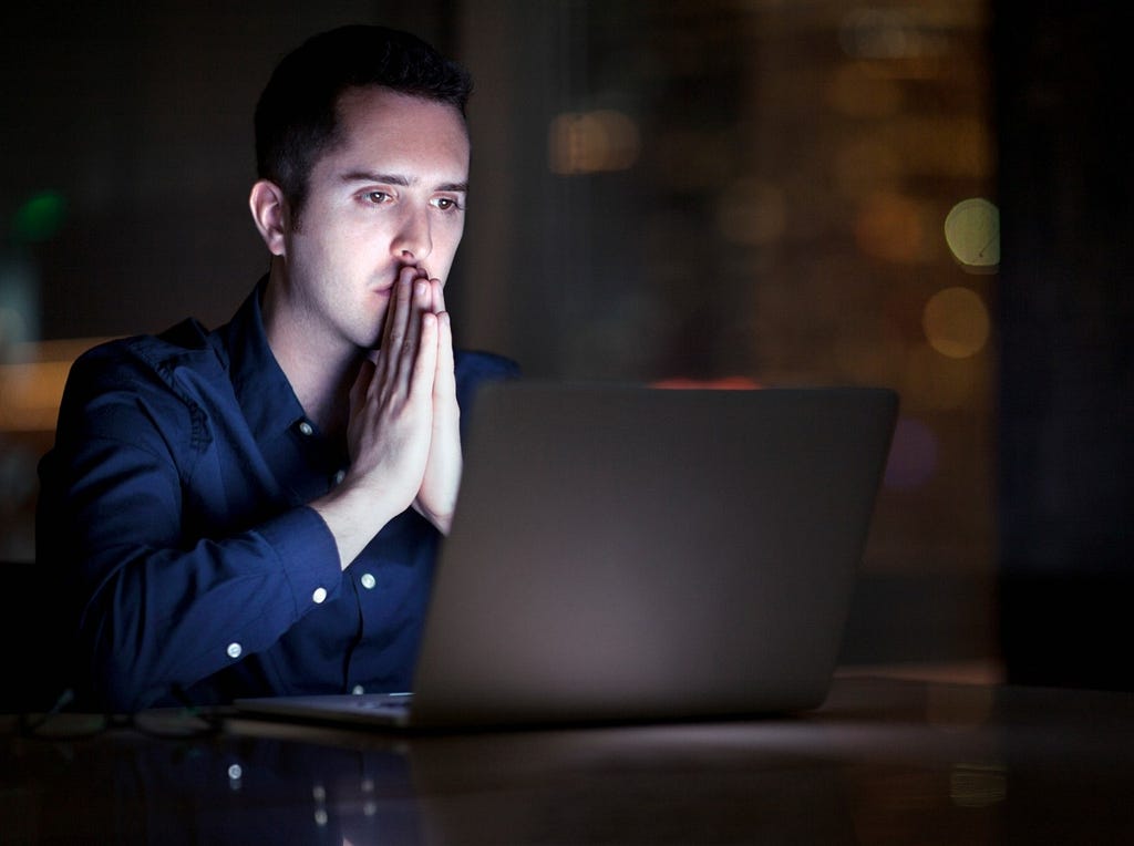 A white man sitting in a dimly lit room with a laptop on the table in front of him and his hands together in front of his mouth. Photo by Shannon Fagan/Getty Images