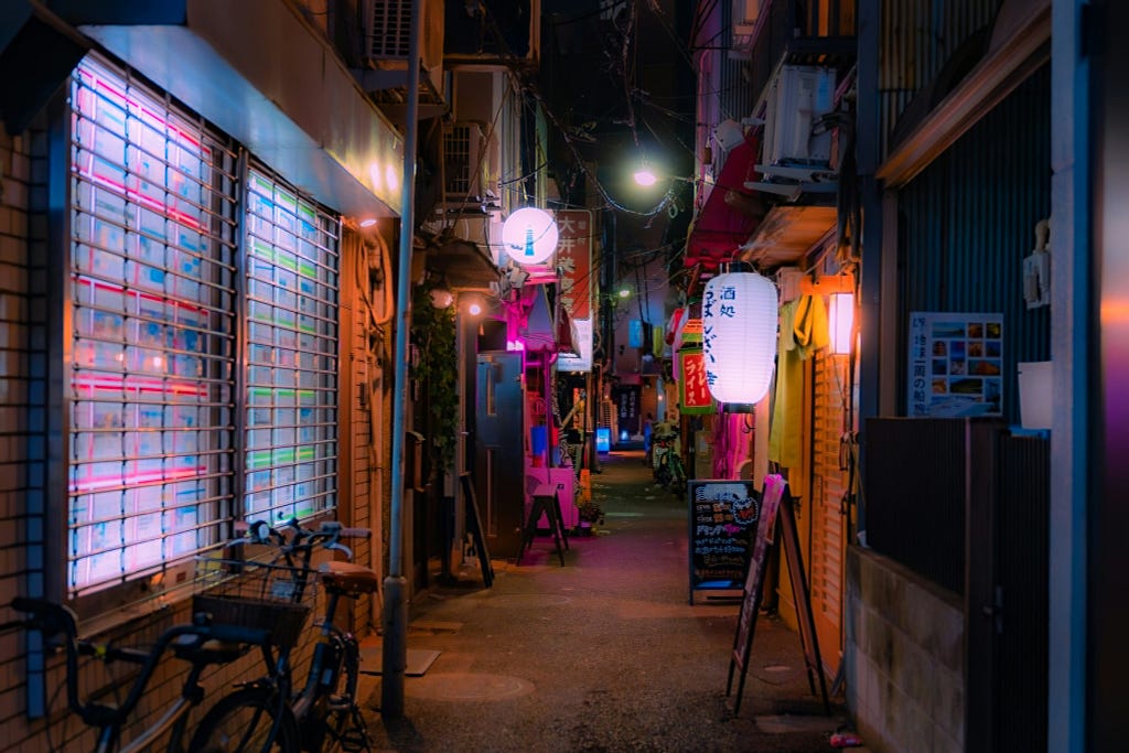 A narrow alley in a city at night, illuminated by the warm glow of paper lanterns and neon lights. Bicycles are parked beside a building on the left, and small, cozy eateries with signboards are scattered along the alley. Overhead cables crisscross above, adding to the urban tapestry of the scene.
