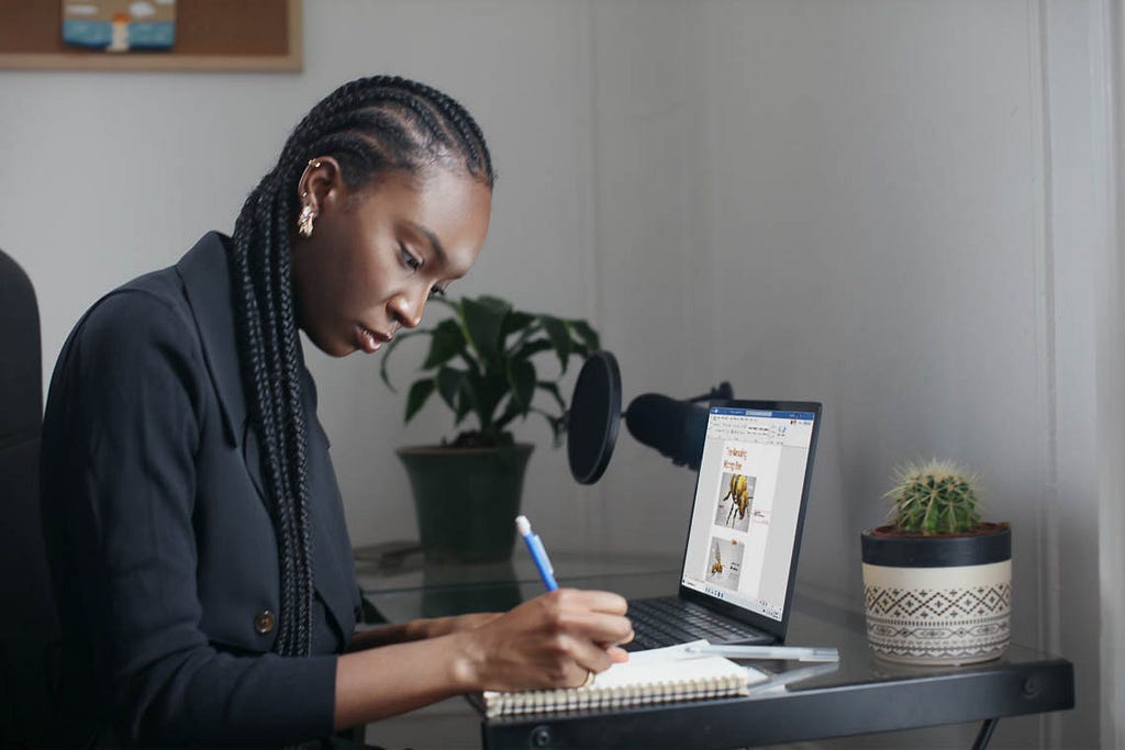 Black woman writing in a notebook beside a laptop