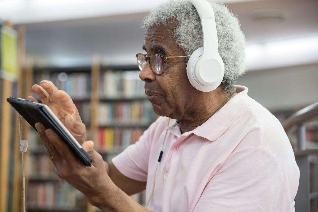 An elderly man with white hair and dark complexion wearing earphones navigate his fingers across an electronic tablet