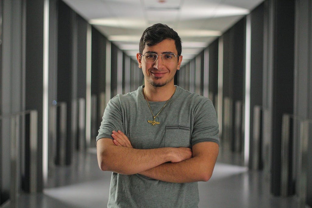 A portrait photograph of Shahan wearing glasses and a green t-shirt, in front of a long well-illuminated hallway.