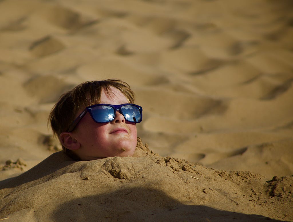 Kid wearing sunglasses is buried in sand up to his neck.