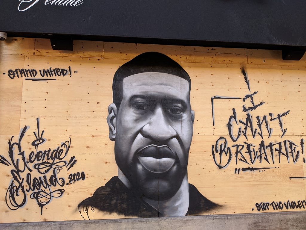 Grey-scale mural of George Floyd on a plywood covered storefront with the words “I Can’t Breathe!” “Stand United” and “Stop the Violence”