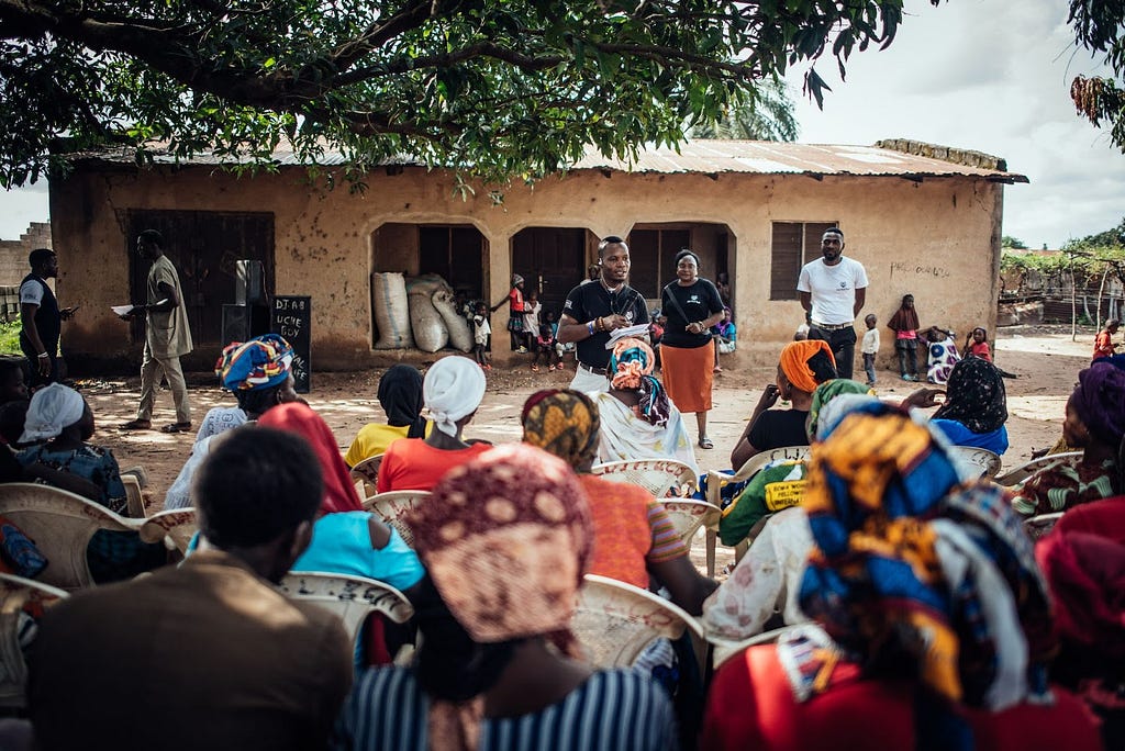 A young man speaks into a microphone at an outdoor community forum, in a rural setting in Nigeria. Several community members are seated in rows, facing the speaker and with their backs to the camera.