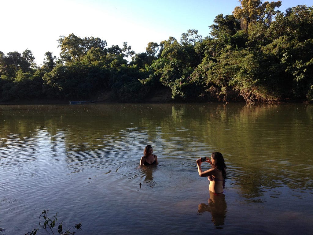 É fim de tarde. No fundo, há uma mata e, diante dela, um rio de águas esverdeadas. Duas moças estão no rio e uma fotografa a outra.