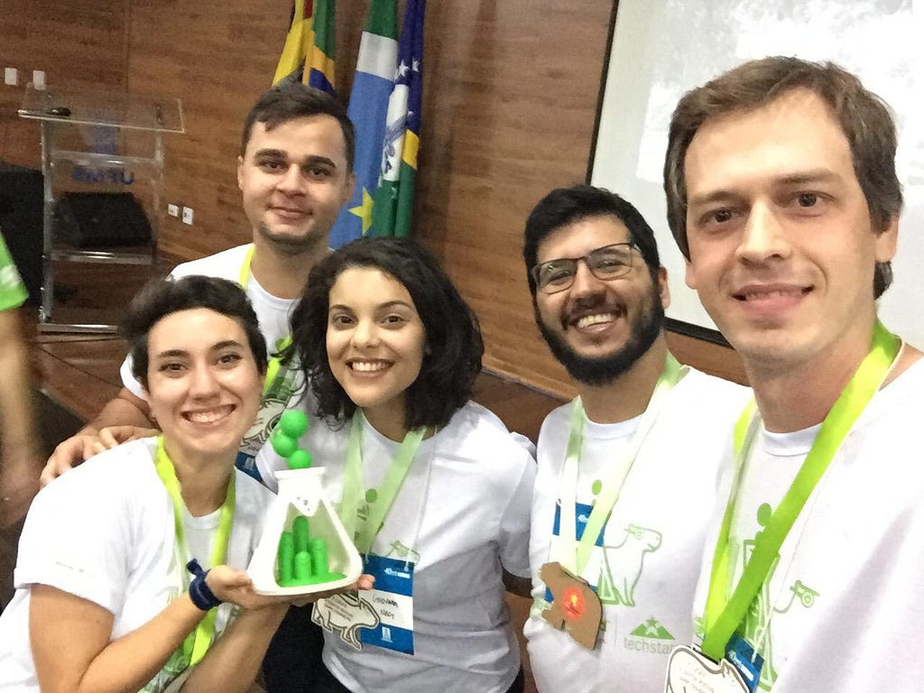 Cinco pessoas sorrindo e se abraçando. Uma delas segura um trófeu de primeiro lugar