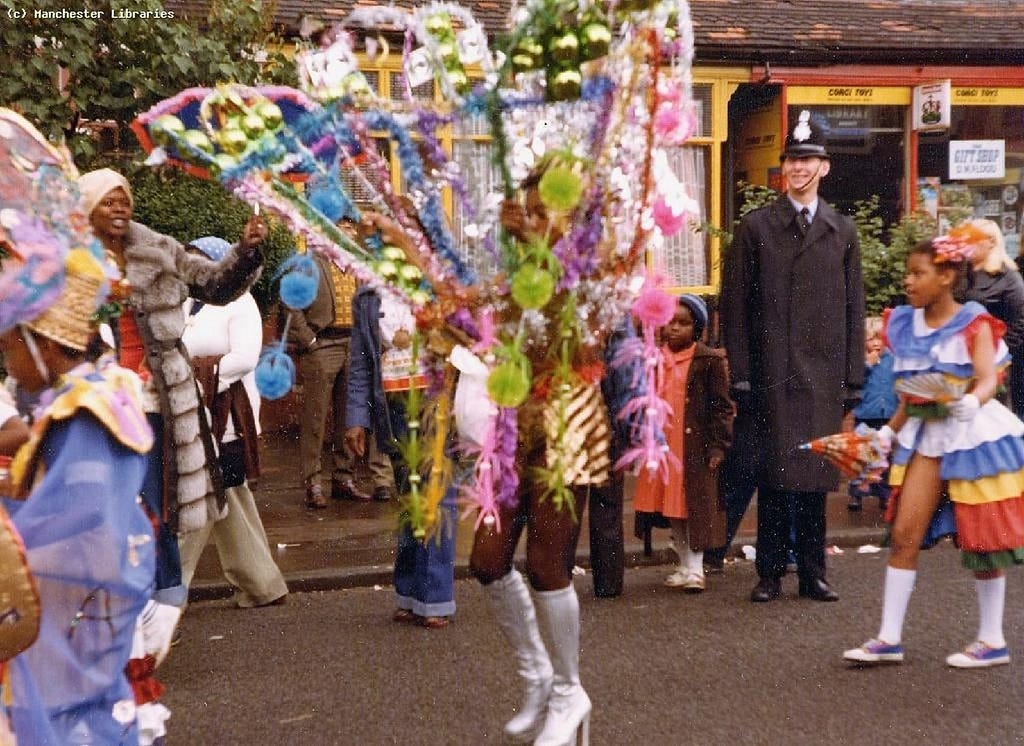 Photograph: Moss Side Carnival dancers in elaborate dress parade along the street. A policeman stands right of the frame watching on and smiling with other onlookers.