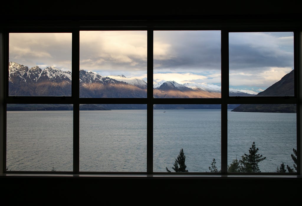 view of lake, snowy mountains, and sun-lit clouds through a multi-paned window