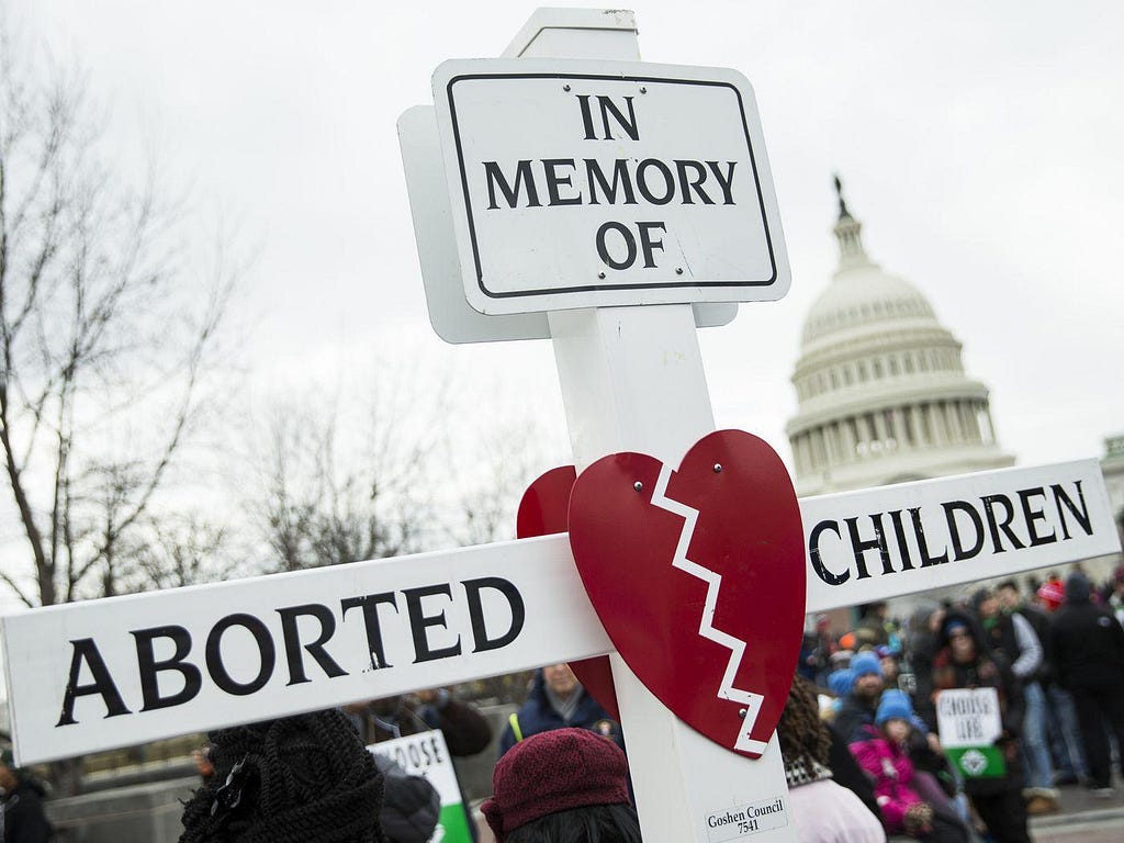 An anti-abortion sign in the shape of a cross. The cross is white, with a broken red heart at its center. The words “In memory of aborted children” is written across the cross’s vertical and horizontal beams.