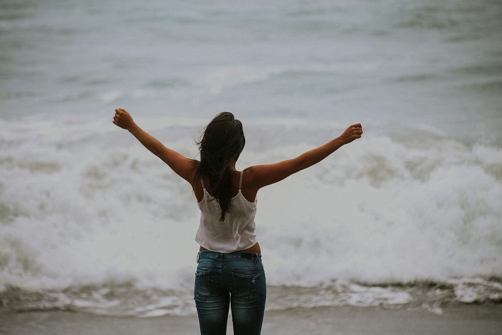 An audacious woman outstretches her arms as a sudsy wave approaches her at the beach on an overcast day.