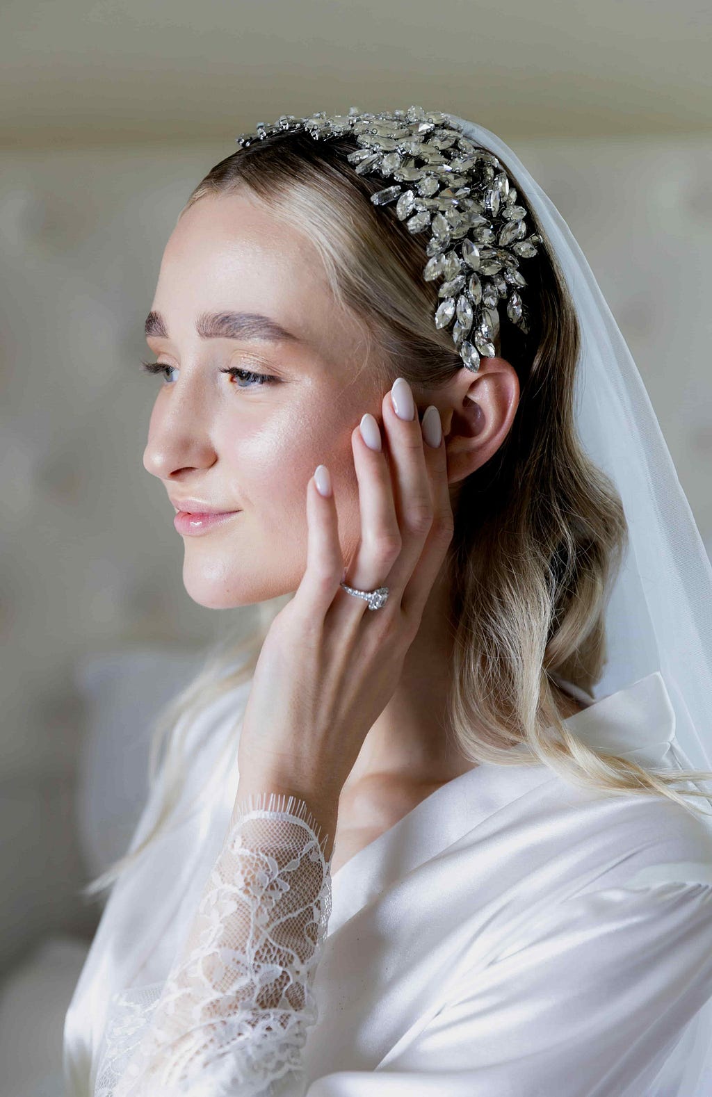 Close-up of a bride wearing a custom engagement ring with man-made diamonds. She is gently touching her face, showcasing the intricate details of her jeweled headpiece and veil, along with her elegant lace sleeve.