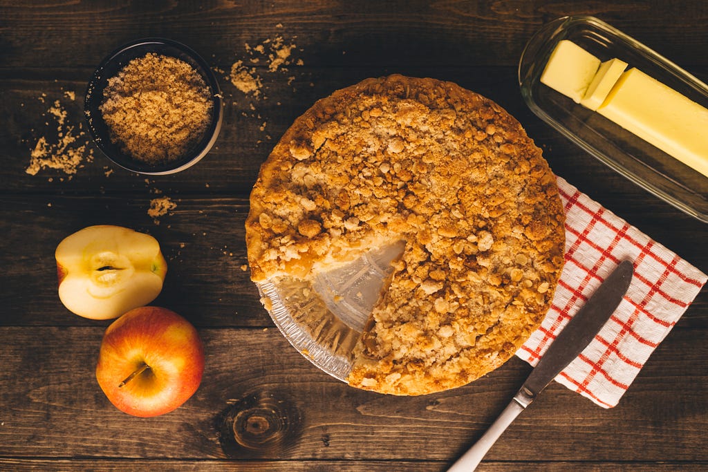 An apple crumble with a slice cut out which is nowhere to be seen. Next to it there’s a block of butter, 1 whole apple, 1 less than half and apple, extra crumble, because who doesn’t love some crumble and a knife set upon a white with red stripes tea towel.