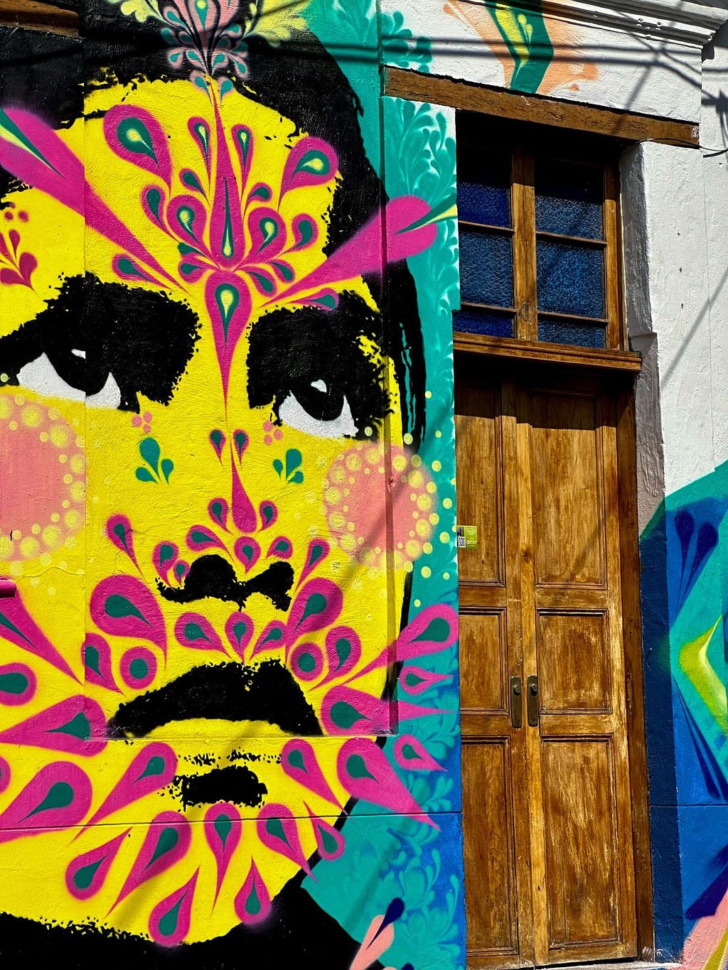 A vibrant mural of a face with intricate pink and yellow patterns covers a wall next to a wooden door, under a blue sky.