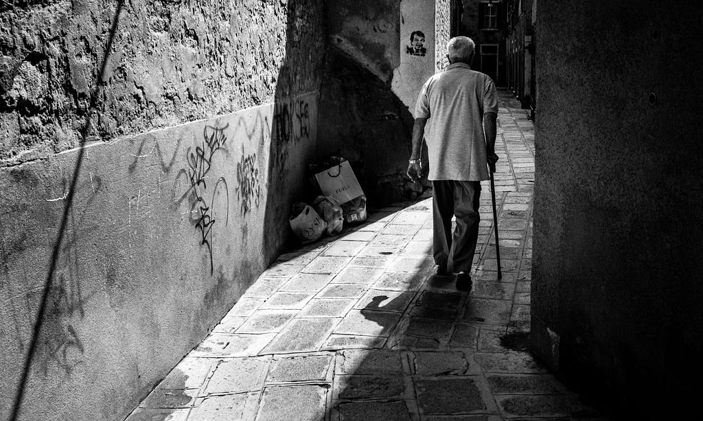 A sad black-and-white image of a lonely old man with stick, with his back turned, who is walking through an empty narrow alley, heading to the unknown.