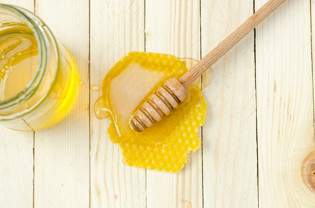 A honey dipper in a pool of honey on a wood paneled surface sits next to a glass jar of that same honey