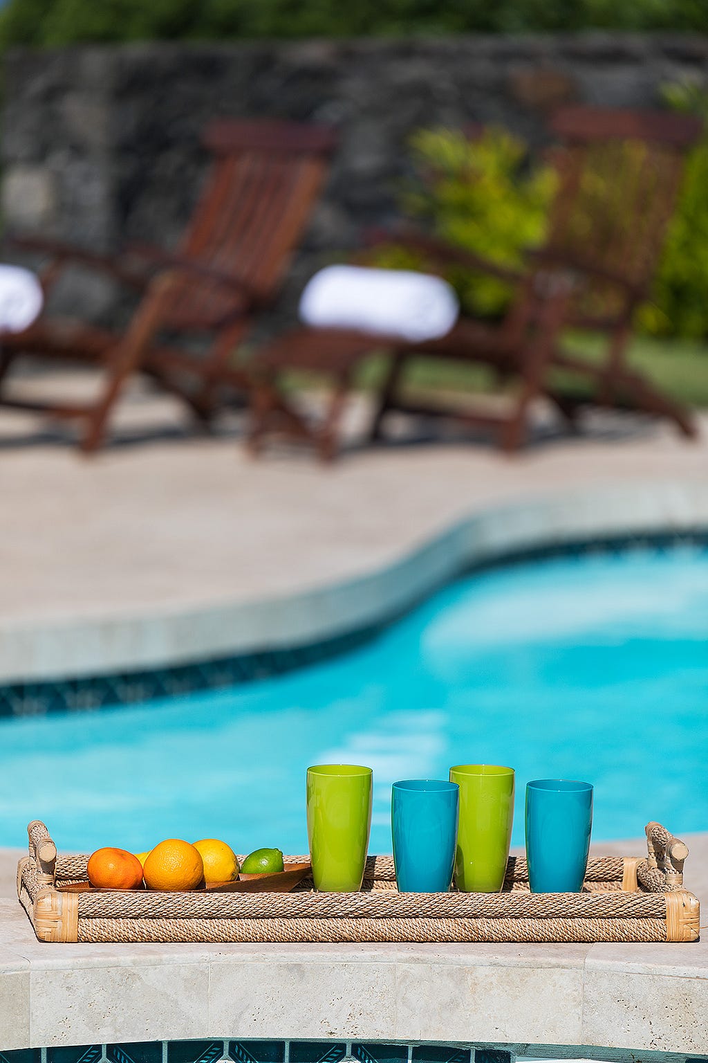 Pool in the background. A woven tray displaying green plastic glasses and fruit in the foreground.