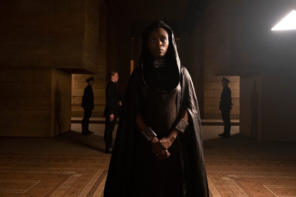 This is a still image from the movie Dune (2021). The scene has brutalist beige and brown architecture wich geometric patterns on the ground. A Black woman with a black hooded cape and textured gown stands in the front. Her right hand clasps her left. She has a necklace with many rings that coverns her neck. There are three soldiers, two men with black uniforms and caps and a woman soldier with her hair in the bun in the background. There is a light in the background into the left.