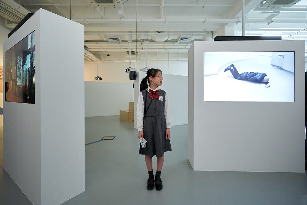 A young student speaks inside a gallery space next to two plinths displaying video works. She is looking towards her left.