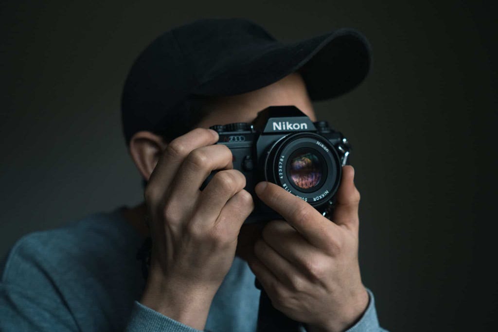 Young man wearing hat holding an old style Nikon camera up to his face to take a photo