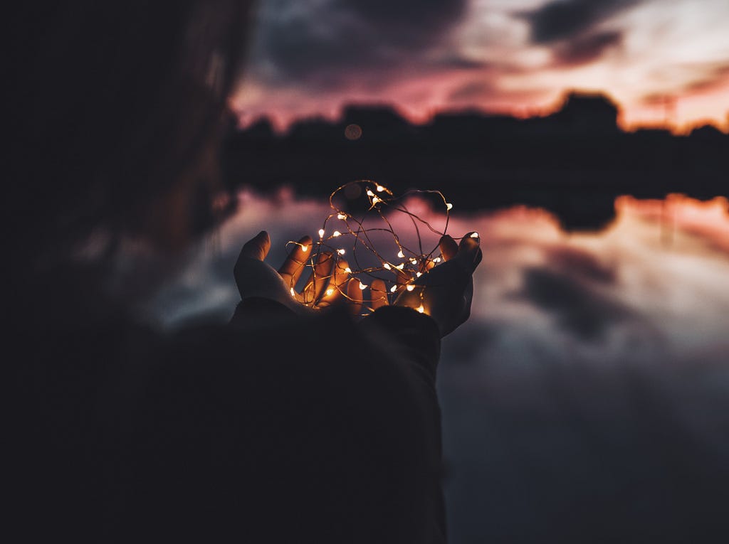 A picture of hands holding out a string of glittering lights in the backdrop of a dark horizon with red colors in the surrounding sky. It depicts one setting themselves apart so they can glow to attract the same glow from however far it is.
