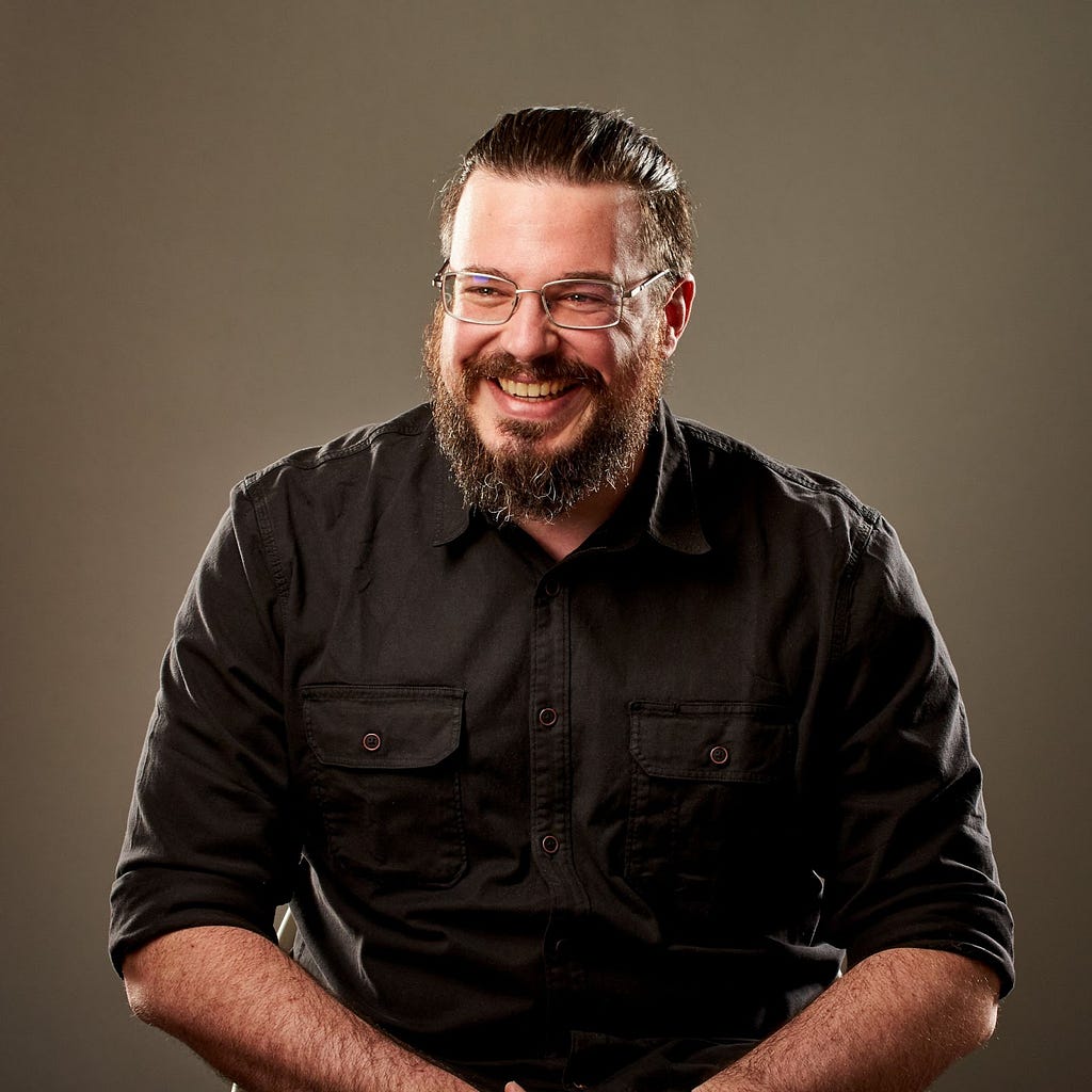Justin Kenny’s headshot — He sits in front of a beige background and appears to be mid-laugh. He’s has a beard and is wearing glasses and a black button-down shirt.