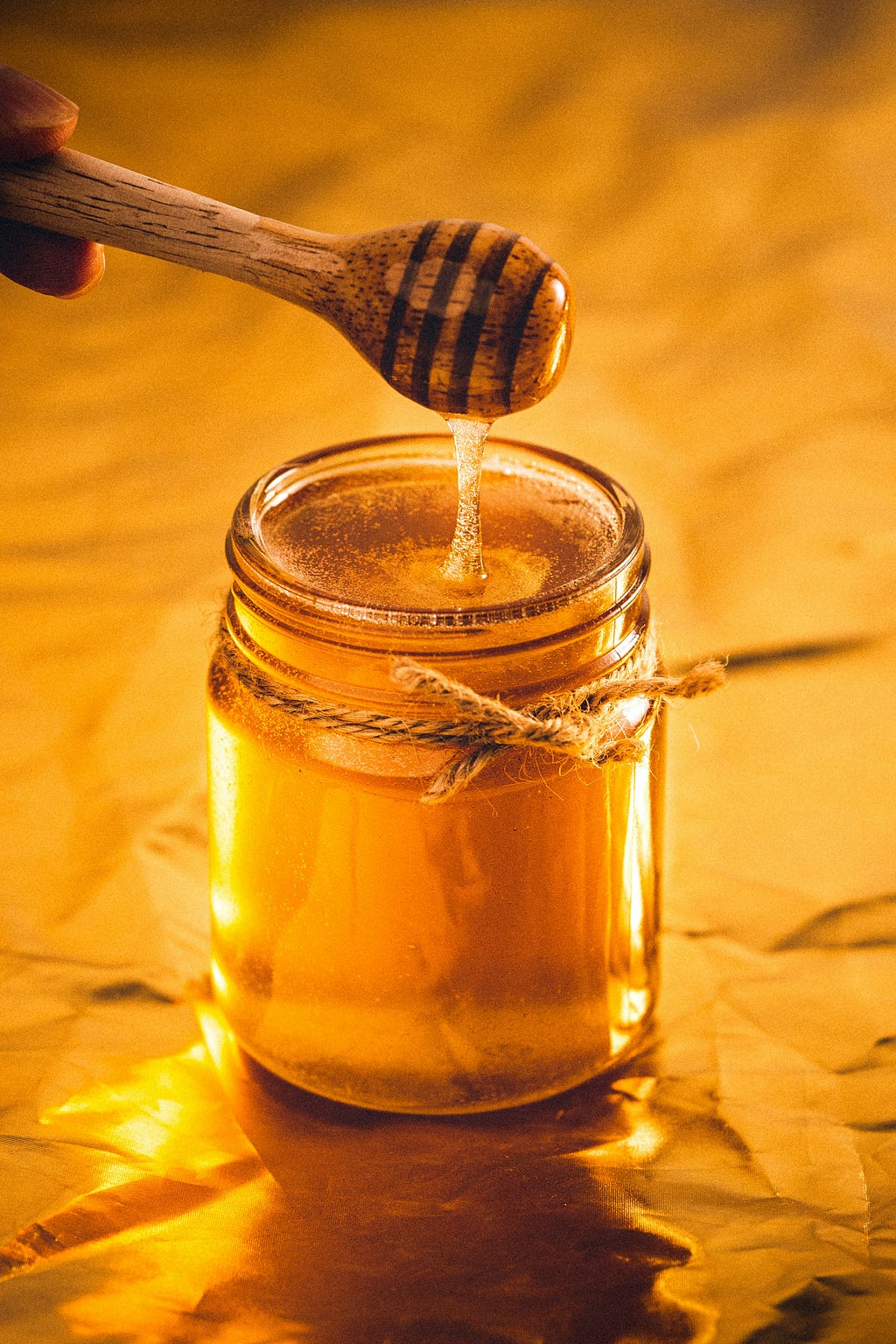 Yellow background. A big Jar full of honey. A honey dipper on top of the jar dripping honey.